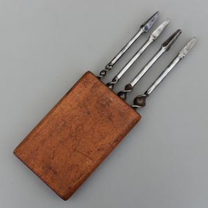 Może przedstawiać: Zestaw czterech metalowych narzędzi z ostrymi końcówkami i skręconymi uchwytami, przechowywanych w prostokątnym drewnianym etui. Narzędzia są srebrne, a etui brązowe z widocznym usłojeniem i zadrapaniami.