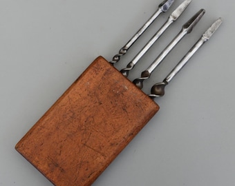 May include: A set of four metal tools with pointed tips and twisted handles, stored in a rectangular wooden case. The tools are silver-colored, and the case is a warm brown with visible grain and scratches.