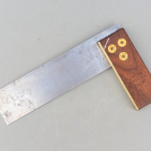 May include: A vintage metal and wood try square with brass accents. The square has three brass rivets on the wooden handle. The metal blade is slightly tarnished.