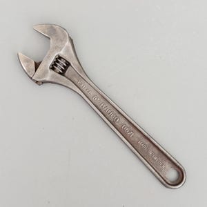 May include: An adjustable wrench, marked "V-STEEL 8" BAHCO 0671 MADE IN SWEDEN", lies on a gray surface. The wrench is silver-colored and has an adjustable jaw. The tool is approximately 8 inches long.