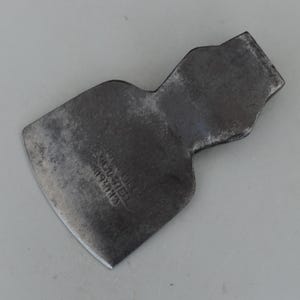 May include: A vintage, dark gray axe head with a slightly curved blade. The metal shows signs of wear and age, with visible text that reads "TOOL STEEL" and "PAT. 192013". The axe head has a rectangular shape with a curved cutting edge.