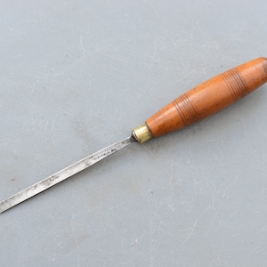 Puede incluir: Un cincel con mango de madera y hoja de metal. La hoja está marcada con "Manufactured by The Stanley Rule & Level Co. New Britain, Conn. U.S.A."