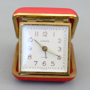 Puede incluir: Un reloj de viaje rojo y dorado vintage con esfera blanca. La esfera del reloj presenta números negros, manecillas doradas y la palabra "EUROPA". El reloj está abierto, revelando la esfera y las palabras "2 JEWELS" y "GERMANY".