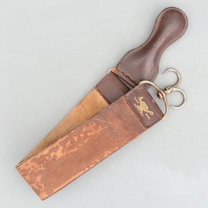 Puede incluir: Un cuero de afeitar de cuero marrón con un asa y herrajes metálicos. El cuero tiene un respaldo de lona y una superficie de cuero desgastada. El texto "TRADE ROMA BRITISH MARK" es visible. Una herramienta de afeitado clásica.