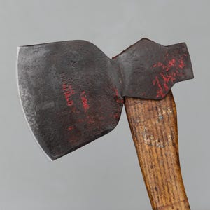 May include: Close-up of a vintage axe with a dark, weathered steel blade and a wooden handle. The blade has red markings and the handle shows signs of wear. The axe is designed for chopping wood and other outdoor tasks.