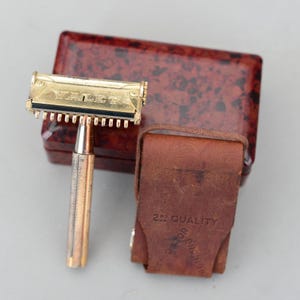 Könnte beinhalten: Ein Vintage-Rasierapparat-Set in Goldoptik mit dem Wort "VALET" auf dem Rasiererkopf. Das Set enthält ein braunes Lederetui und eine gemusterte, rotbraune Box. Das Lederetui ist mit den Worten "2nd QUALITY RAZOR ENGLAND" versehen.