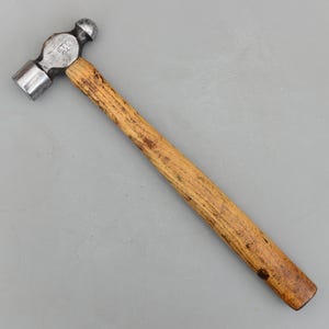 Puede incluir: Un martillo de bola con mango de madera y cabeza de acero. La cabeza del martillo tiene una cara plana por un lado y una pena redondeada en forma de bola por el otro. El mango es de madera marrón claro.