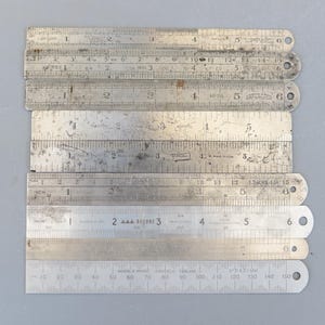 Op de afbeelding: Een verzameling vintage metalen linialen, elk met inch-metingen. De zilverkleurige linialen vertonen tekenen van slijtage, met duidelijke markeringen en cijfers. Sommige linialen hebben een gat aan het einde. De langste is ongeveer 38 cm.