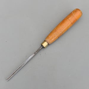 Puede incluir: Un formón para trabajar la madera con mango de madera marrón claro y hoja de metal. El mango tiene ranuras horizontales. Una virola de latón une el mango a la hoja de metal. La hoja es plateada y termina en punta.