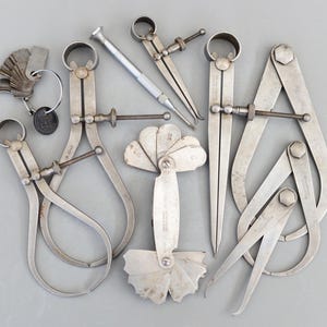 Puede incluir: Una colección de herramientas de medición metálicas antiguas, que incluyen calibradores, un destornillador y un juego de galgas. Las herramientas son de color plateado y están dispuestas sobre una superficie gris. Los calibradores varían en tamaño y diseño.