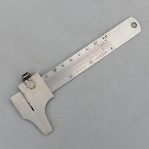 Peut inclure: Un pied à coulisse métallique vintage avec une échelle coulissante, marquée de chiffres et de mesures en pouces. L'outil possède un mécanisme coulissant et une petite boucle à l'extrémité. Le pied à coulisse est conçu pour une mesure précise. (environ 25.4 cm)