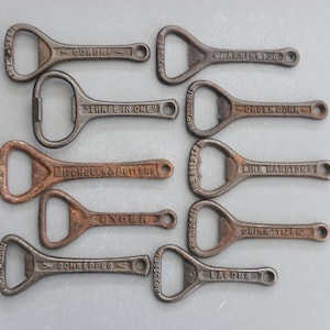 Może przedstawiać: Kolekcja dziesięciu zabytkowych otwieraczy do butelek z różnymi nazwami marek, w tym Corona, Three in One, Mitchells & Butlers, Gyder, Schweppes, Lacons, GHarrington, Crown Cork, Like Marstonst i Drink Mitzer.