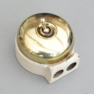 May include: A vintage-style light switch with a brass-colored, domed cover and a small toggle. The switch is mounted on a cream-colored base with two holes. The design is reminiscent of older electrical fixtures.
