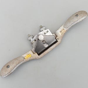 May include: A vintage Stanley No. 80 cabinet scraper, a hand tool for woodworking. The tool has a silver-colored body with textured handles and a black blade. It features adjustment screws and a yellow label.