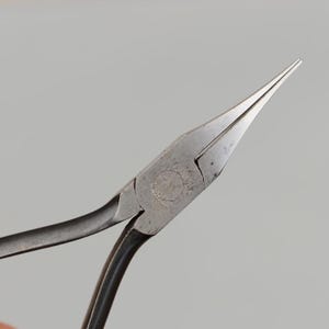 May include: Close-up of needle-nose pliers, a hand tool with long, tapered jaws. The pliers are made of metal with a dark, slightly worn finish. The handles curve smoothly, and the jaws are pointed and precise.