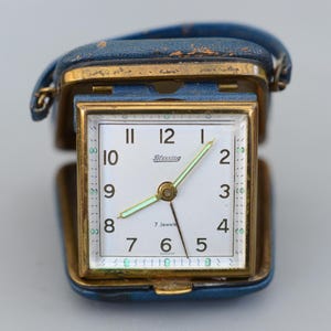 May include: A vintage travel alarm clock in a blue leather case. The clock face is white with black numerals and the brand name "Blessing". The hands are gold and the case is rectangular with a gold frame. The clock is open, revealing the face.