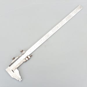 May include: A vintage silver-coloured caliper tool with measurement markings. The tool has a sliding jaw and a long, graduated beam for precise measurements. The caliper is designed for measuring the internal or external dimensions of an object.