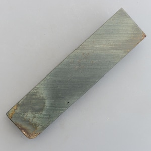 May include: A rectangular sharpening stone, approximately 15 cm long, with a grey-green colour. The stone has a textured surface with visible striations and a slightly worn edge. It is a tool for sharpening blades.