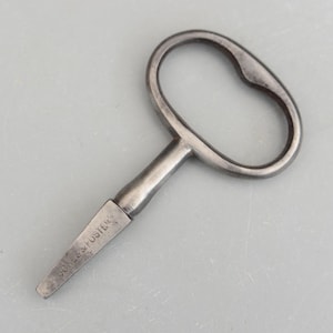 以下が含まれることがあります： 楕円形のハンドルと長方形のビットが付いた、銀色の金属製のキー。キーのビットには「JONES & FOSTER」の刻印があります。このヴィンテージキーは、時計やその他の機械装置用である可能性があります。