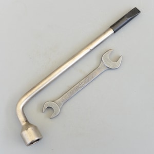 Puede incluir: Dos herramientas metálicas: una llave de rueda y una llave inglesa. La llave de rueda es plateada con un mango negro. La llave inglesa es plateada y tiene el texto "14 NISSAN MOTOR 17" impreso. Ambas herramientas están sobre una superficie gris.