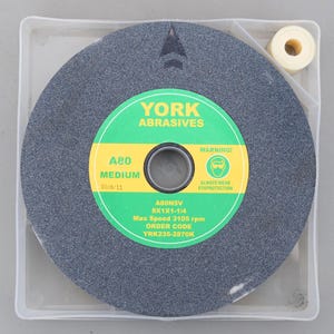 Op de afbeelding: Een grijze slijpschijf met een groen en geel etiket met de tekst "YORK ABRASIVES". Het etiket bevat ook de tekst "A80 MEDIUM", "8X1X1-1/4" en "Max Speed 3105 rpm". Een kleine rol witte tape staat in de hoek.