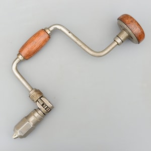 May include: A vintage hand drill with a wooden handle and a metal crank. The drill is silver with a brass-coloured crank.