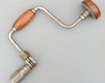 Op de afbeelding: Een vintage handboor met een houten handvat en een metalen kruk. De boor is zilverkleurig met een messingkleurige kruk.