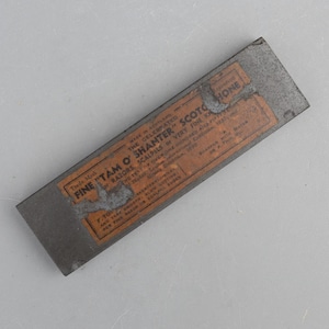 Könnte beinhalten: Ein rechteckiger grauer Schleifstein mit einem verwitterten orangefarbenen Papieretikett und schwarzem Text, der Fine Tam O' Shanter Scotch Hone, Made in Scotland, for razors, scalpels, or very fine knives und sharpen with water or a thin oil lautet.