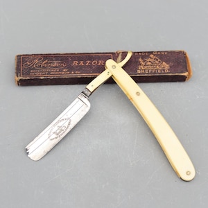 Könnte beinhalten: Ein Vintage-Rasiermesser mit einem weißen Knochengriff und einer silbernen Klinge. Die Klinge ist mit dem Text "Manufactured by Herbert Robinson & Co. Cutlery Works Sheffield" graviert. Das Rasiermesser befindet sich in seiner Originalverpackung, die braun mit goldenen Lettern ist. Die Schachtel trägt den Text "Robinson Razor" und "Trade Mark".