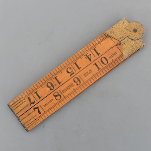 Peut inclure: Une règle pliante en bois de couleur orange avec des marquages noirs et des chiffres, dont "Rabone" et "Made in England". La règle mesure jusqu'à 43 cm et possède des raccords en laiton.