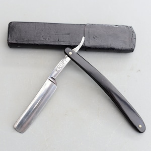 May include: A vintage straight razor with a black handle and a silver blade. The blade is inscribed with the word "KROPP". The razor is in a black leather case.