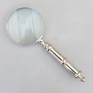 Peut inclure: Une loupe argentée avec une lentille ronde et une poignée décorative. La poignée présente une série d'anneaux et une extrémité pointue. La lentille est claire, et la loupe est posée sur un fond gris clair.
