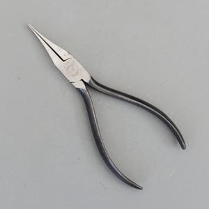 May include: A pair of needle-nose pliers with a silver head and black handles. The pliers are designed with long, tapered jaws for precision work. The handles are curved for a comfortable grip. The tool is set against a gray background.