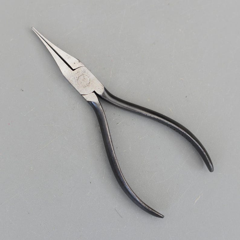 May include: A pair of needle-nose pliers with a silver head and black handles. The pliers are designed with long, tapered jaws for precision work. The handles are curved for a comfortable grip. The tool is set against a gray background.