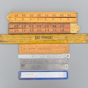 Puede incluir: Una colección de reglas antiguas de varios tamaños y materiales, incluyendo madera y metal. Algunas reglas son plegables y otras rectas. Una regla tiene el texto "GRE-SOLVENT" y "Made its mark by taking 'em off!! The Elbow Grease is in the Tin!"