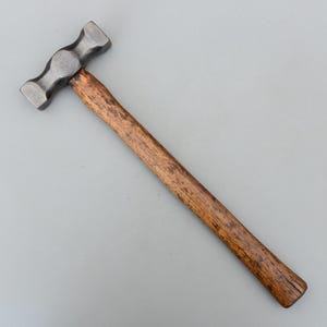 Peut inclure: Marteau en métal avec une tête sombre et texturée et un long manche en bois. La tête du marteau a une forme arrondie avec des surfaces de frappe plates. Le manche est de couleur marron chaud, montrant le grain naturel du bois.