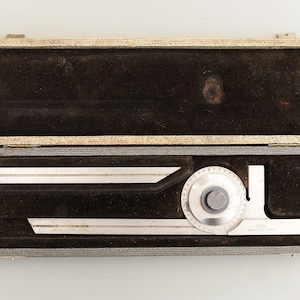 Może przedstawiać: Srebrny kątomierz z obracającym się tarczą i linijką w szarym, aksamitnym etui. Kątomierz jest oznaczony w stopniach i ma mały otwór w środku tarczy.