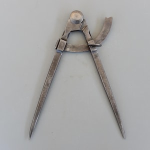 Puede incluir: Un compás de metal vintage con dos patas puntiagudas y un brazo curvo. El compás es de color plateado y parece estar hecho de acero. La herramienta está diseñada para medir y dibujar círculos. El compás está sobre un fondo gris.
