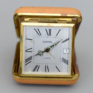 Puede incluir: Un reloj de viaje Europa vintage en un estuche de cuero marrón. La esfera del reloj es plateada con números romanos y manecillas negras. El reloj tiene un marco dorado y se muestra la fecha. Las palabras "EUROPA" y "2 JEWELS" están impresas en la esfera.