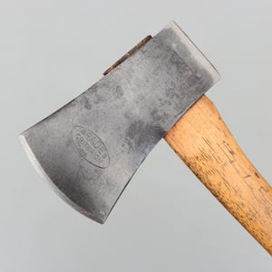 May include: Close-up of a vintage axe with a wooden handle and a steel blade. The blade has the text "BRADES CRITERION 1212" stamped on it. The axe head is a dark gray color, and the handle is light brown.
