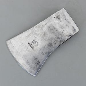 May include: A silver-colored axe head with a curved blade and a flat back. The axe head has a worn, textured appearance and the text "SWEDEN DROP FORGED" is stamped on the surface. The axe head is designed for chopping wood.