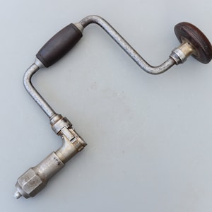Peut inclure: Une perceuse à main vintage avec une poignée et un bouton en bois. La perceuse est en métal et présente un design incurvé. La poignée est marron foncé et le bouton est marron clair. La perceuse est conçue pour un fonctionnement manuel.