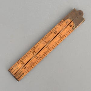 May include: A vintage folding wooden ruler featuring two hinged sections with black printed markings and numbers. The light brown wood displays visible grain, while the metal hinge mechanism at the top is dark brown with a circular pivot point.