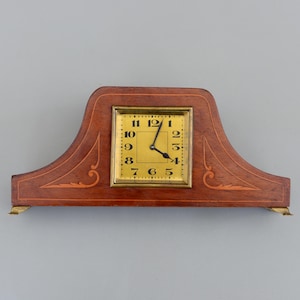Puede incluir: Un reloj de sobremesa de madera vintage con una esfera rectangular dorada y números negros. La estructura de madera del reloj es marrón con incrustaciones decorativas y patas de latón. Las manecillas del reloj señalan aproximadamente las 4:58. Mide unos 25 cm de largo.