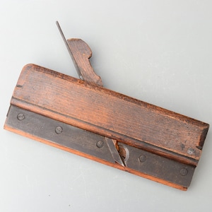 May include: A vintage wooden hand plane with a metal blade and a wooden handle. The plane is made of dark wood and has a worn finish. It has several screws on the metal blade.