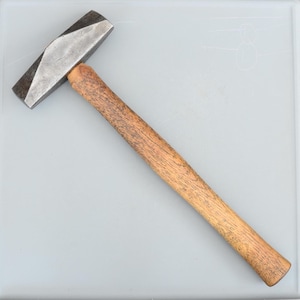 Puede incluir: Un martillo de metal con mango de madera. La cabeza del martillo es de color gris oscuro y tiene un diseño facetado. El mango es de madera marrón claro.