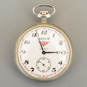 Può includere: Un orologio da tasca argentato con quadrante bianco, numeri neri e dettagli rossi. Il quadrante dell'orologio presenta il marchio "SERKISOF" e il testo "16 WELS" e "MADE IN USSR". L'orologio ha un piccolo quadrante dei secondi e un anello per catena argentato.
