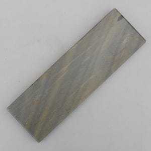 Könnte beinhalten: Ein rechteckiger Schleifstein mit flacher Oberfläche, der eine gesprenkelte grau-braune Farbpalette mit subtilen, diagonalen Streifenmustern auf der gesamten Vorderseite aufweist. Der Stein hat scharfe, saubere Kanten und eine gleichmäßige, dichte Textur.