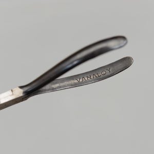 May include: Close-up of a metal tool with curved arms. The tool has a dark, matte finish and the word "VANALOY" is stamped on one of the arms. The tool appears to be used for gripping or manipulating small objects.