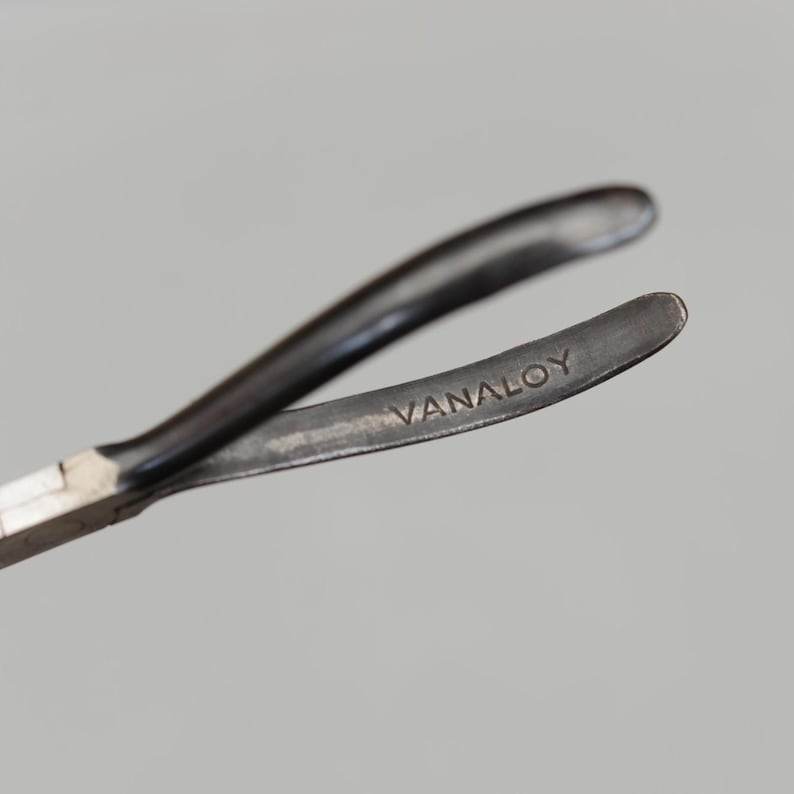 May include: Close-up of a metal tool with curved arms. The tool has a dark, matte finish and the word "VANALOY" is stamped on one of the arms. The tool appears to be used for gripping or manipulating small objects.